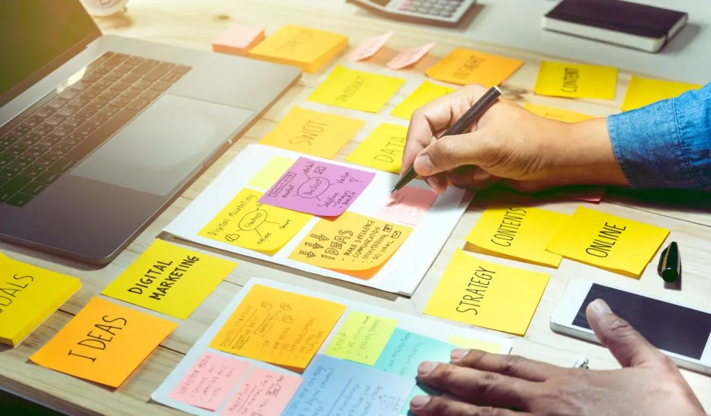 Manos escribiendo notas adhesivas de marketing digital y estrategia en mesa de trabajo