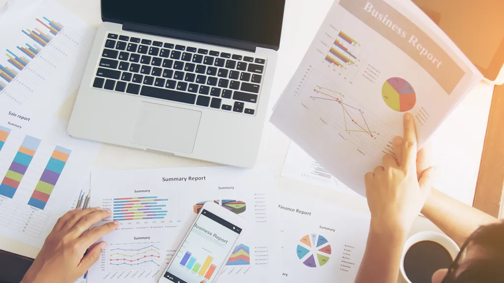 Manos revisando informes de negocio con gráficos en laptop, smartphone y documentos en escritorio