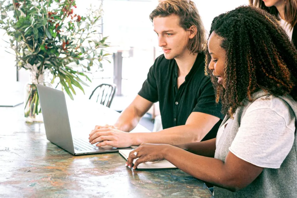Dos personas colaborando con un laptop y cuaderno, planta decorativa en el fondo