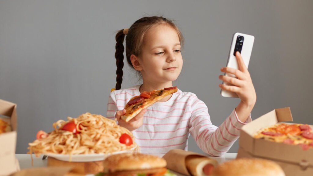 Niña sentada en una mesa con comida chatarra viendo en un teléfono celular publicidad inadecuada sobre alimentos.